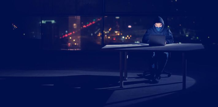 Young talented hacker using laptop computer while working in dark office with big city lights in the background at night