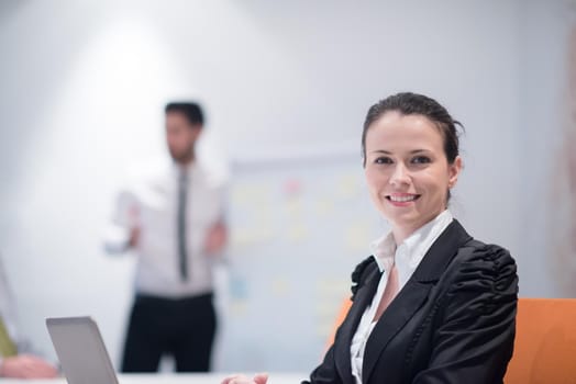 young business woman on meeting usineg laptop computer, blured group of people in background at  modern bright startup office interior taking notes on white flip board and brainstorming about plans and ideas