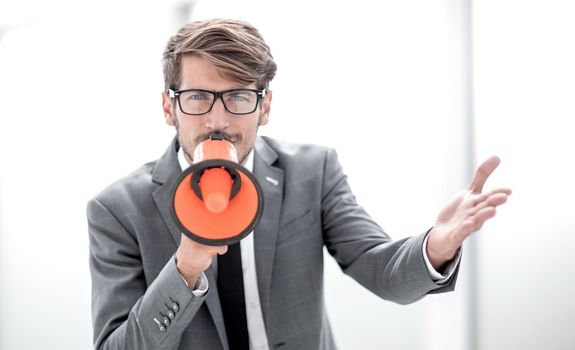 Portrait of furious business man screaming with megaphone in modern office. The angry man looks at camera. Copy space