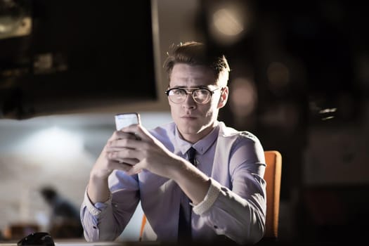 Young man using mobile phone while working on computer at night in dark office.