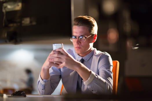 Young man using mobile phone while working on computer at night in dark office.