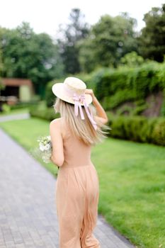 Back view of young blonde woman in body color overalls and hat standing in yeard. Concept of fashion and summer season.