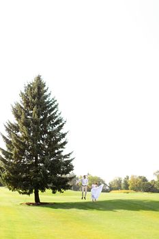 Young couple walking near green big spruce on grass in white sky background. Concept of evergreens and wedding.