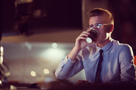 Tired businessman working late doing overtime in office at night drinking coffee to go on.