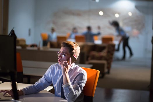 Young man using mobile phone while working on computer at night in dark office.