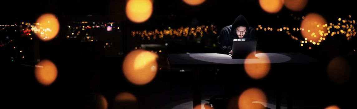 Young talented hacker using laptop computer while working in dark office with big city lights in the background at night