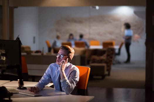 Young man using mobile phone while working on computer at night in dark office.