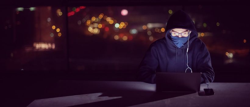 Young talented hacker using laptop computer while working in dark office with big city lights in the background at night