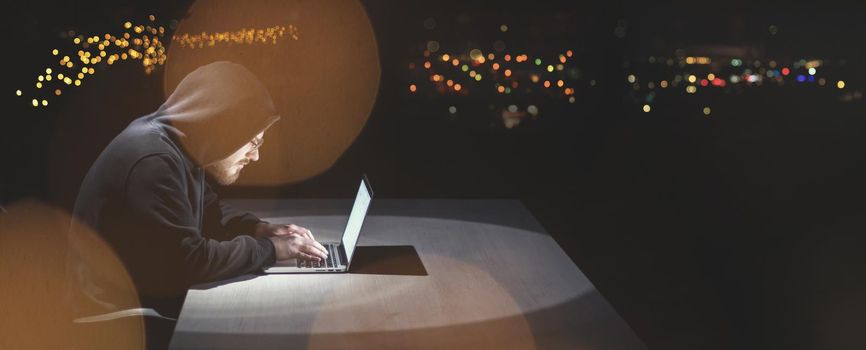 Young talented hacker using laptop computer while working in dark office with big city lights in the background at night