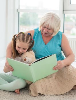 Cute granny reading book to little granddaughter holding toy sitting on floor in playroom