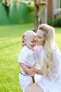 Young blonde mother playing and sitting with little kid on grass in yard and kissing son. Concept of motherhood and child.