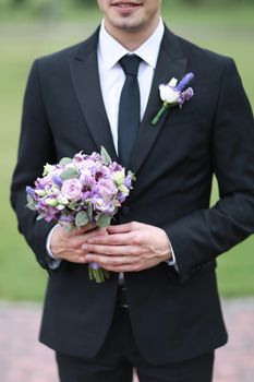 Groom wearing black suit and tie keeping bouquet of flowers. Concept of wedding.