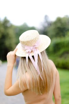 Back view of young blonde girl in body color overalls and hat standing in yeard. Concept of fashion and summer season.