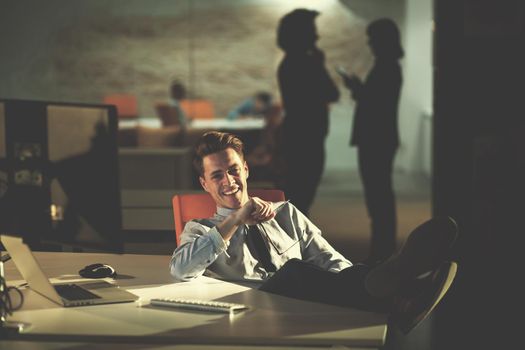 Full length of a relaxed casual young businessman sitting with legs on desk at night office