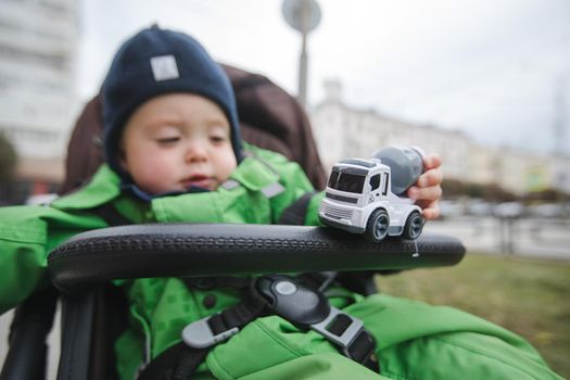 Adorable infant in warm clothing sitting in stroller and playing with toy car outside.