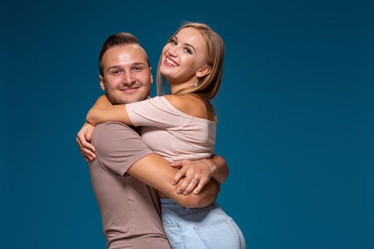 Young couple is hugging on blue background in studio. They wear T-shirts, jeans and smile. Friendship, love and relationships concept