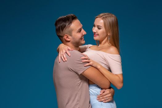 Young couple is hugging on blue background in studio. They wear T-shirts, jeans and smile. Friendship, love and relationships concept