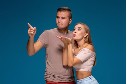 Portrait of handsome man and beautiful woman or young couple looking away with funny face and kissing with love. Indoor studio shot, isolated on blue background.
