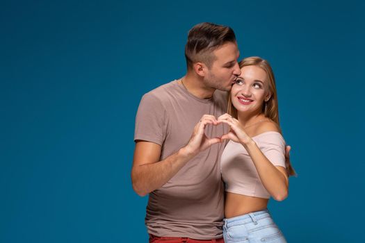 Happy couple dressed casual clothes, making heart shape from fingers, studio portrait on blue background. Friendship, love and relationships concept