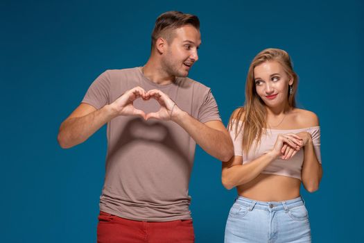 Happy couple dressed casual clothes, making heart shape from fingers, studio portrait on blue background. Friendship, love and relationships concept