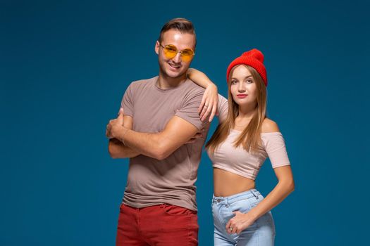 Studio lifestyle portrait of two best friends hipster wearing stylish bright outfits, hats, denim shorts and glasses, going crazy and having great time together. Indoor studio shot, isolated on blue background.