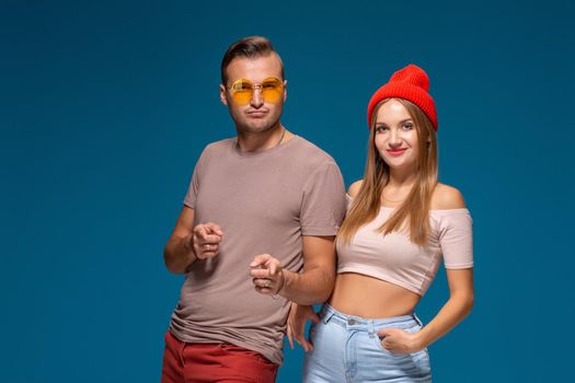 Studio lifestyle portrait of two best friends hipster wearing stylish bright outfits, hats, denim shorts and glasses, going crazy and having great time together. Indoor studio shot, isolated on blue background.