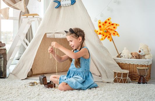 Little girl exploring jewellery from small chest sitting on room's floor in front of wigwam