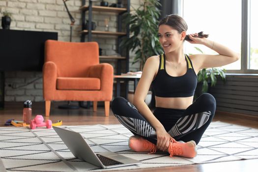 Fitness beautiful slim woman is sitting on the floor with dumbbells and bottle of water using laptop at home in the living room. Stay at home activities