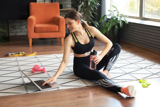 Fitness beautiful slim woman is sitting on the floor with dumbbells and bottle of water using laptop at home in the living room. Stay at home activities