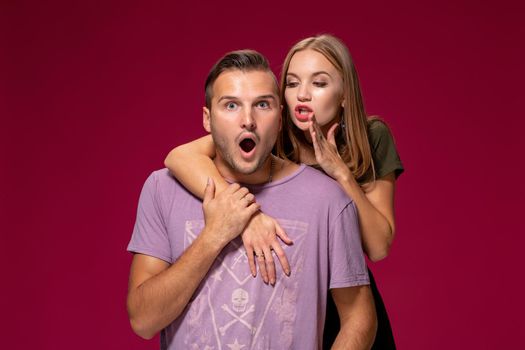 Studio shot of secret young woman and man keep index fingers on lips, stand next to each other, tell private information, ask not spread rumors and be quiet, isolated over burgundy background.