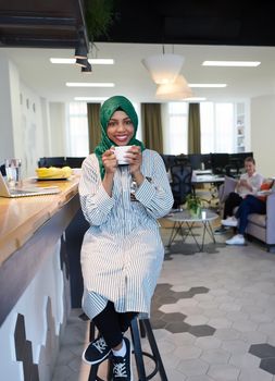 african muslim business woman wearing a green hijab drinking tea while working on laptop computer in relaxation area at modern open plan startup office. Diversity, multiracial concept