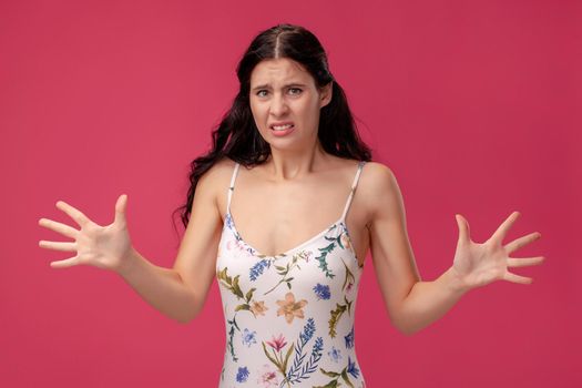 Portrait of a pretty woman in a white dress with floral print standing on a pink wall background in studio. She gestures something big and unpleasant. People sincere emotions, lifestyle concept. Mockup copy space.