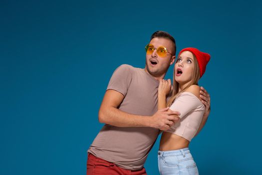 Surprised couple in casual clothes pointing with their index fingers up looking there with great astonishment isolated over blue background with copy space for your advertisement or written text.