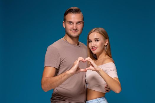 Happy couple dressed casual clothes, making heart shape from fingers, studio portrait on blue background. Friendship, love and relationships concept