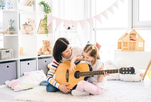 Pretty mother teaching cute daughter to play guitar sitting on floor in children's room