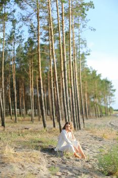 Young girl sitting on sand beach with trees in backgroundand wearing white clothes. Concept of freedom and summer vacations