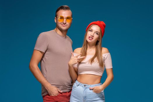 Studio lifestyle portrait of two best friends hipster wearing stylish bright outfits, hats, denim shorts and glasses, going crazy and having great time together. Indoor studio shot, isolated on blue background.