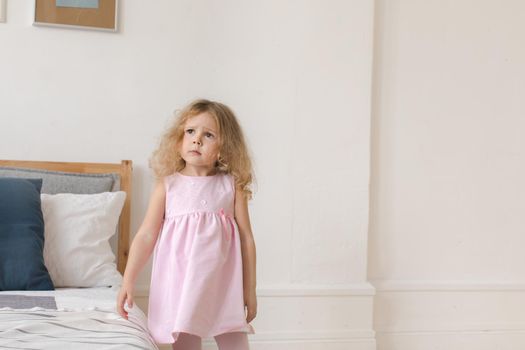 Tender little girl in pink dress standing in cozy bedroom and looking away