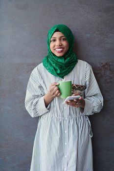 african muslim businesswoman with green hijab using mobile phone during coffee break from work in front of gray wall outside