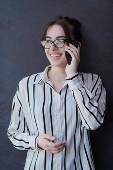 startup businesswoman in shirt with a glasses using mobile phone while standing in front of gray wall during break from work outside