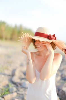 Young cute woman in hat with red ribbon standing on shingle beach. Concept of summer vacations and resort.