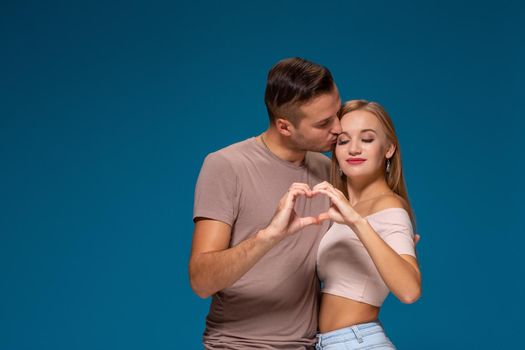 Happy couple dressed casual clothes, making heart shape from fingers, studio portrait on blue background. Friendship, love and relationships concept