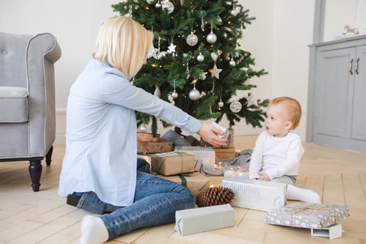 Side view of young blond woman sitting with little child on parquet near Christmas decorated tree among wrapped gifts