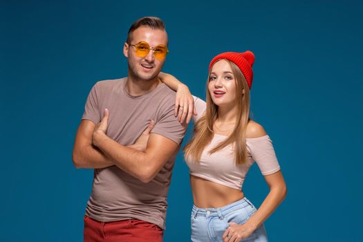 Studio lifestyle portrait of two best friends hipster wearing stylish bright outfits, hats, denim shorts and glasses, going crazy and having great time together. Indoor studio shot, isolated on blue background.