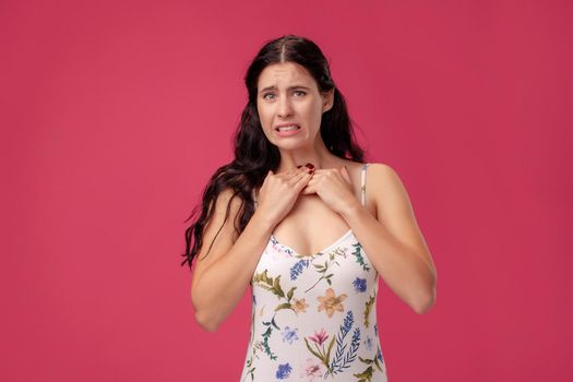 Portrait of a pretty young woman in a white dress with floral print standing on a pink wall background in studio. She is having a sore throat and holding her neck. People sincere emotions, lifestyle concept. Mockup copy space.