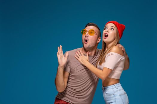 Surprised couple in casual clothes pointing with their index fingers up looking there with great astonishment isolated over blue background with copy space for your advertisement or written text.