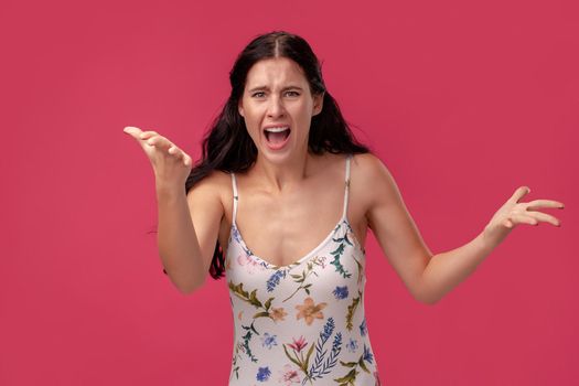 Portrait of a woman in a white dress with floral print standing on a pink wall background in studio. She swears at someone and screams loudly. People sincere emotions, lifestyle concept. Mockup copy space.