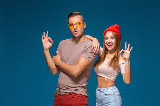 Joyful couple in casual wear smiling and gesturing ok sign isolated over blue background. Studio lifestyle portrait of two best friends hipster