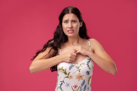 Portrait of a young woman in a white dress with floral print standing on a pink wall background in studio. She is very worried about something. People sincere emotions, lifestyle concept. Mockup copy space.