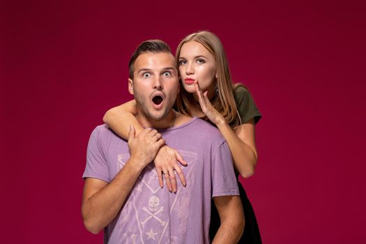 Studio shot of secret young woman and man keep index fingers on lips, stand next to each other, tell private information, ask not spread rumors and be quiet, isolated over burgundy background.